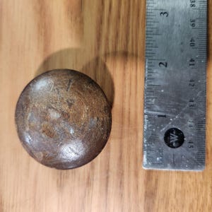 May include: A circular, dark brown wooden knob with a smooth, polished surface. The knob is positioned next to a metal ruler that shows the object has a diameter of approximately 1.5 inches. The wood grain is visible across the surface.
