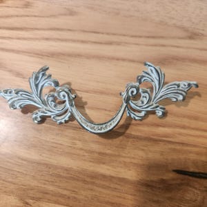 May include: A decorative metal drawer pull featuring an ornate, symmetrical scrollwork design. The hardware has a distressed finish with a white painted surface over a dark metallic base, highlighting the intricate curves and leaf-like patterns.