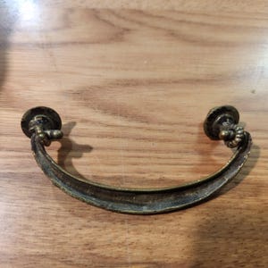 May include: A curved, antique brass drawer pull featuring a weathered, dark patina finish. The handle is attached to two circular, textured mounting plates with small, rope-like detailing at the connection points, creating a classic, functional hardware piece.
