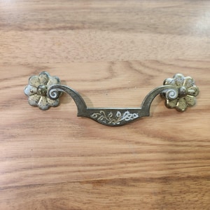 May include: A metal drawer pull featuring a curved handle with an engraved leaf pattern and two circular flower-shaped backplates. The hardware has a weathered, tarnished brass finish with visible dark spots and light-colored highlights.