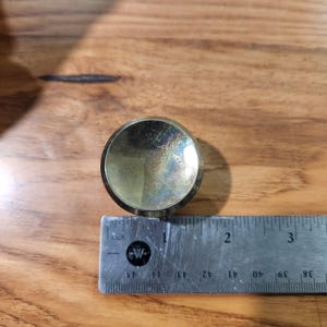 May include: A circular, metallic gold-toned knob with a reflective, slightly iridescent surface. The knob is positioned directly above a metal ruler, showing a diameter of approximately 1.25 inches across its widest point.