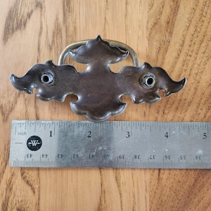 May include: A dark metal drawer pull with a curved handle and an ornate backplate featuring two mounting posts. A metal ruler is positioned underneath, showing the hardware measures approximately 4.5 inches in total width.