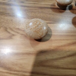 May include: A round cabinet knob with a smooth, glossy finish in a light beige and tan marbled pattern. The knob features a domed top surface and a cylindrical base, showcasing a subtle, mottled texture throughout the light-colored material.