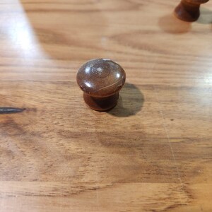 May include: A small, round, brown wooden knob with a smooth, polished finish. The top surface features a subtle, light-colored circular marking and a faint, thin scratch across the center, showing a natural wood grain texture throughout the piece.
