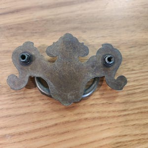 May include: A brass-toned metal drawer pull featuring a decorative backplate with an ornate, symmetrical shape and a curved, swinging handle. The backplate includes two threaded metal posts for mounting, showing a weathered, aged patina finish.