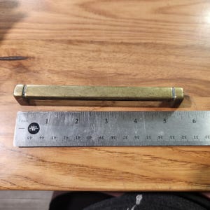 May include: A rectangular brass cabinet pull with a weathered finish, positioned above a metal ruler. The pull measures approximately 5.5 inches in total length, featuring a straight bar design with two mounting points at the ends.