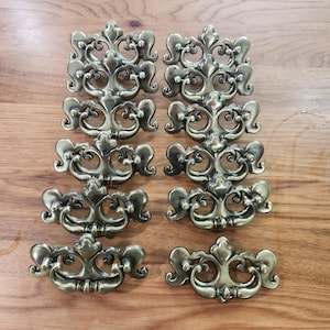 May include: A set of ten brass-toned metal drawer pulls arranged in two vertical columns. Each pull features a symmetrical, ornate scrollwork design with a central fleur-de-lis motif and two mounting points for attachment to furniture.