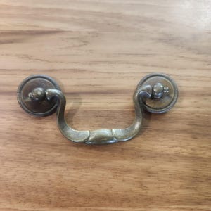 May include: A brass-toned metal drawer pull featuring a curved, swinging handle attached to two circular backplates. Each backplate is decorated with a rope-like textured border and a central knob, providing a secure mounting point for furniture.