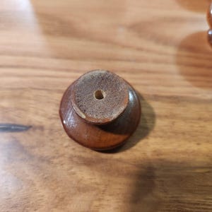 May include: A small, round, brown wooden knob with a smooth, polished finish on its curved exterior. The flat top surface is unfinished and textured, featuring a single, centered circular hole for mounting purposes.
