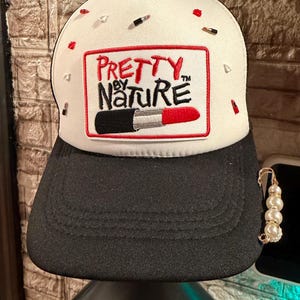 May include: A white and black trucker hat featuring a rectangular patch with the text PRETTY BY NATURE and a lipstick graphic. The white front panel is decorated with small red and black lipstick-shaped charms, and a gold safety pin with pearls.