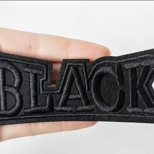 May include: A black embroidered patch featuring the word BLACK in a bold, block-style font. The letters are stitched with a raised texture onto a matching black fabric base, creating a cohesive and monochromatic appearance for the entire piece.
