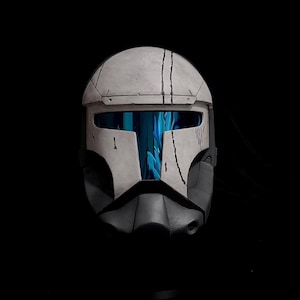 May include: A white and dark gray tactical helmet featuring a T-shaped visor with a reflective blue tint. The helmet surface displays weathered black scratch marks and panel lines, creating a battle-worn appearance against a solid black background.
