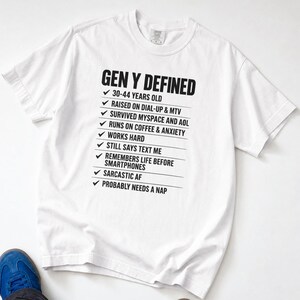 May include: A white short-sleeved t-shirt featuring black text that reads GEN Y DEFINED followed by a checklist of items including 30-44 YEARS OLD, RAISED ON DIAL-UP & MTV, SURVIVED MYSPACE AND AOL, RUNS ON COFFEE & ANXIETY, WORKS HARD, STILL SAYS TEXT ME.