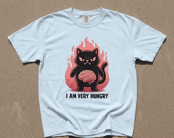 Puede incluir: Una camiseta de manga corta azul claro con un gráfico de un gato negro con un cerebro rosa en el estómago, rodeado de llamas rojas. Debajo del gato, el texto dice I AM VERY HUNGRY en letras mayúsculas negras. Tiene un cuello redondo.