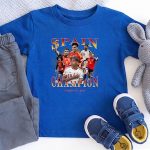 May include: A royal blue short-sleeve t-shirt featuring a graphic print of the Spanish national soccer team players. The text reads SPAIN CHAMPION WORLD CUP 2023 in a yellow and red gradient font. The design includes multiple players in red jerseys.