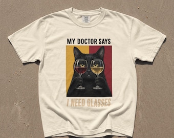 Puede incluir: Una camiseta de manga corta color crema con un gráfico de un gato negro sosteniendo dos copas de vino. El texto superior dice MY DOCTOR SAYS y el inferior I NEED GLASSES. El fondo detrás del gato está dividido en secciones amarillas y rojas.