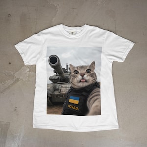 Peut inclure: Un t-shirt blanc à manches courtes avec une impression graphique représentant un chat tigré portant un gilet tactique noir avec un écusson aux couleurs bleu et jaune et le texte "УКРАЇНА" en jaune, devant un char militaire sous un ciel nuageux.