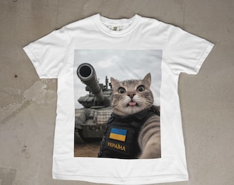 Puede incluir: Una camiseta blanca de manga corta con un estampado gráfico de un gato atigrado que lleva un chaleco táctico negro con un parche de bandera azul y amarilla y el texto "УКРАЇНА" en amarillo, frente a un tanque militar bajo un cielo nublado.