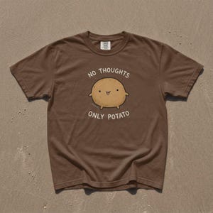 Könnte beinhalten: Ein braunes T-Shirt mit kurzen Ärmeln, das eine Grafik einer beigen Kartoffel mit einem kleinen Gesicht zeigt. Darüber und darunter steht der weiße Text "NO THOUGHTS ONLY POTATO". Das Shirt hat einen Rundhalsausschnitt und eine normale Passform.