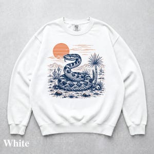 May include: A white crewneck sweatshirt featuring a dark blue graphic illustration of a coiled rattlesnake in a desert landscape. The design includes a large orange sun, distant mountains, cacti, and rocks, with the word White printed at the bottom.