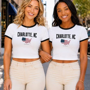 Op de afbeelding: Een wit ringer-crop-topje met zwarte boorden aan de mouwen en de halslijn. Op de voorzijde staat de zwarte tekst CHARLOTTE, NC boven een afbeelding van de kaart van de Verenigde Staten met het Amerikaanse vlagpatroon. Het shirt wordt gedragen met een beige broek.
