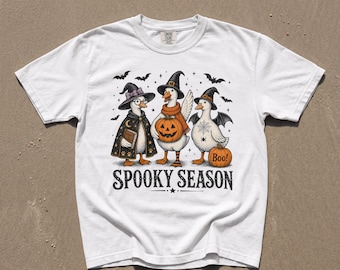 Op de afbeelding: Een wit t-shirt met een afbeelding van drie eenden in Halloween-kostuums, inclusief heksenhoeden, een cape en vleermuisvleugels. Het ontwerp bevat pompoenen, een spinnenweb en vleermuizen, met de tekst SPOOKY SEASON en Boo! eronder gedrukt.