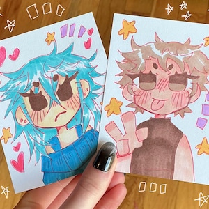 May include: Two rectangular art prints featuring anime-style characters. The left print shows a character with blue hair in a blue sweater, surrounded by pink hearts and yellow stars. The right print shows a character with brown hair making a peace sign.