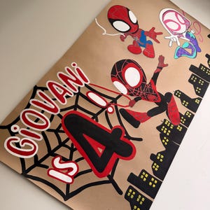 May include: A tan poster featuring hand-painted illustrations of three Spider-Man characters, a black spiderweb, and a black city skyline. The text reads "GIOVANI IS 4" in large red and black lettering, with the number 4 prominently displayed.