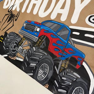 May include: A hand-painted sign on brown paper featuring a blue monster truck with red flame decals, large black tires, and yellow suspension springs. The text "HAPPY BIRTHDAY" is written in large white letters at the top, with a black spider illustration.