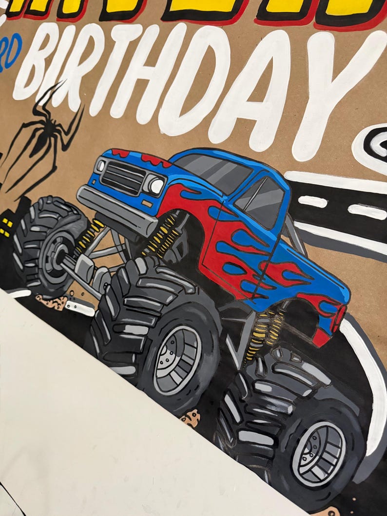 May include: A hand-painted sign on brown paper featuring a blue monster truck with red flame decals, large black tires, and yellow suspension springs. The text "HAPPY BIRTHDAY" is written in large white letters at the top, with a black spider illustration.
