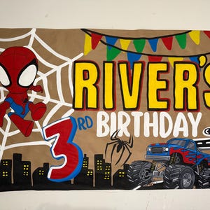 May include: A rectangular birthday banner on a tan background featuring a cartoon spider-themed character, a blue monster truck with flame decals, a black city skyline, a white spider web, and the text RIVER'S 3RD BIRTHDAY with colorful bunting.