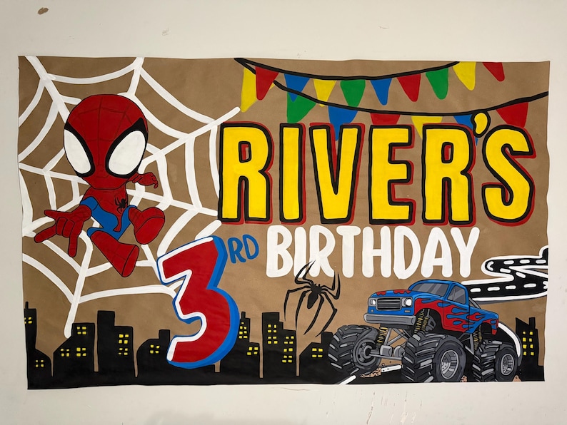 May include: A rectangular birthday banner on a tan background featuring a cartoon spider-themed character, a blue monster truck with flame decals, a black city skyline, a white spider web, and the text RIVER'S 3RD BIRTHDAY with colorful bunting.