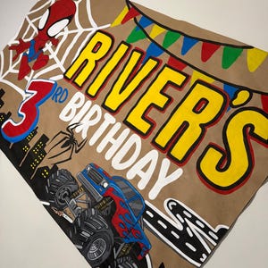 May include: A tan rectangular banner featuring a hand-painted design with a red and blue superhero character, a large red number 3, and a blue and red monster truck. The text reads "RIVER'S 3RD BIRTHDAY" in yellow and white letters.