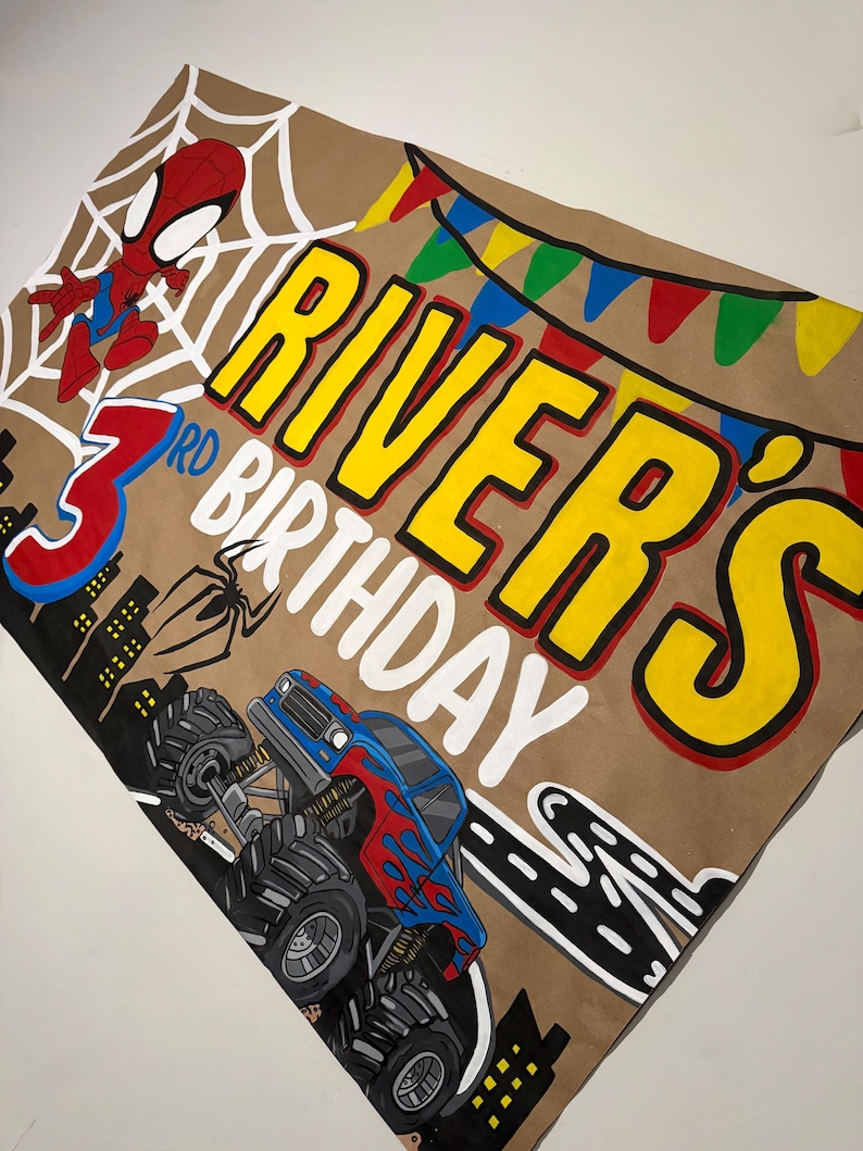 May include: A tan rectangular banner featuring a hand-painted design with a red and blue superhero character, a large red number 3, and a blue and red monster truck. The text reads "RIVER'S 3RD BIRTHDAY" in yellow and white letters.