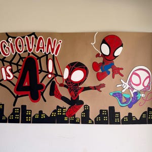May include: A rectangular brown paper poster featuring hand-painted illustrations of three Spider-Man characters in a chibi style. The text reads GIOVANI IS 4! in red and black, accompanied by a black spiderweb, a city skyline, and red webbing.