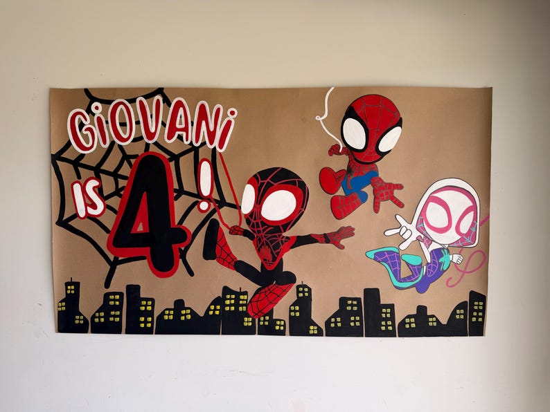 May include: A rectangular brown paper poster featuring hand-painted illustrations of three Spider-Man characters in a chibi style. The text reads GIOVANI IS 4! in red and black, accompanied by a black spiderweb, a city skyline, and red webbing.
