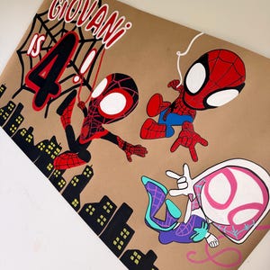 May include: A tan poster featuring the text GIOVANI IS 4, a black spiderweb, and three cartoon superhero characters in red, blue, black, white, and pink. A black silhouette of a city skyline with yellow windows is at the bottom of the poster.