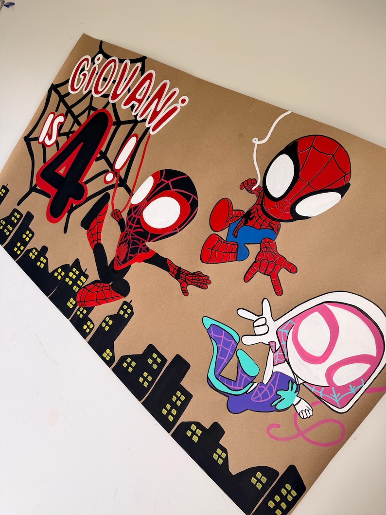 May include: A tan poster featuring the text GIOVANI IS 4, a black spiderweb, and three cartoon superhero characters in red, blue, black, white, and pink. A black silhouette of a city skyline with yellow windows is at the bottom of the poster.