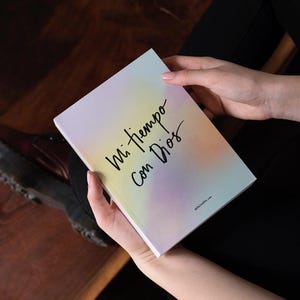 May include: A rectangular book with a soft-focus gradient cover in shades of yellow, orange, and purple. The front cover features the black handwritten text Mi tiempo con Dios and smaller text at the bottom. The book has a clean, white spine.