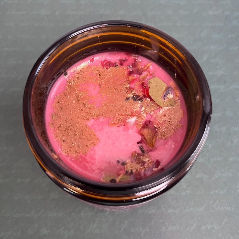 Op de afbeelding: Een bovenaanzicht van een kaars in een donker amberkleurige glazen pot. De was is felroze van kleur, bedekt met een laagje glinsterend bronskleurig poeder en verspreide gedroogde bloemblaadjes. Een enkele witte kaarslont is in het midden zichtbaar.