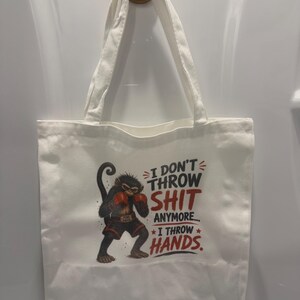 Peut inclure: Un sac fourre-tout en toile blanche avec une illustration de singe portant des gants de boxe rouges et un short noir. Le texte indique : I DON'T THROW SHIT ANYMORE... I THROW HANDS. Le singe a une expression déterminée et une posture de boxeur.