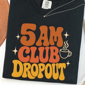 May include: A black crewneck t-shirt featuring the text 5 AM CLUB DROPOUT in large, wavy, retro-style lettering. The words are colored in shades of orange and yellow, accompanied by a small graphic of a steaming coffee cup and four-pointed stars.