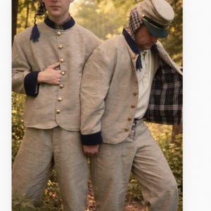 Puede incluir: Dos chaquetas de color beige con cuellos y puños azul marino, con botones dorados en la parte delantera. Una chaqueta está abierta, mostrando un forro de cuadros negros y blancos. Ambas chaquetas se combinan con pantalones beige y una gorra.