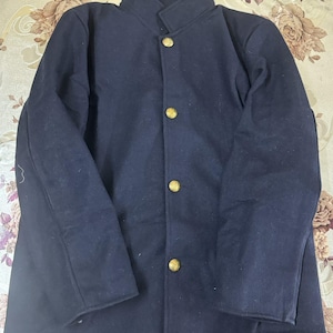 Könnte beinhalten: Eine dunkelmarineblaue Jacke mit hohem Stehkragen und langen Ärmeln. Der vordere Verschluss besteht aus vier runden, goldfarbenen Metallknöpfen, die vertikal angeordnet sind. Das Kleidungsstück besteht aus einem festen, blickdichten Stoff.