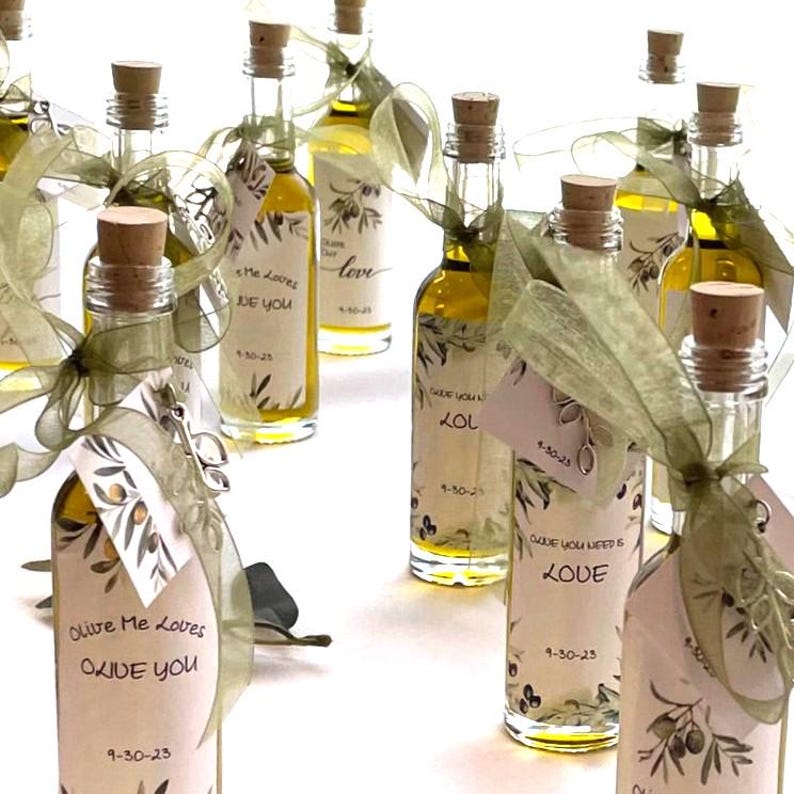 Puede incluir: Peque&ntilde;as botellas de vidrio llenas de aceite de oliva, cada una sellada con un corcho y adornada con una etiqueta y una cinta verde. Las etiquetas presentan ilustraciones de ramas de olivo y frases como "Olive Me Loves Olive You" y "Love".