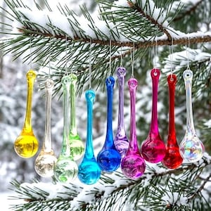 May include: A collection of ten teardrop-shaped glass ornaments hanging from a pine branch. The ornaments feature a smooth, glossy finish in various colors including yellow, clear, green, blue, purple, magenta, red, and an iridescent white finish.
