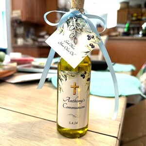 Puede incluir: Una peque&ntilde;a botella de vidrio llena de aceite de oliva, con tap&oacute;n de corcho y etiqueta decorativa. La etiqueta incluye una cruz dorada, el texto "Anthony's Communion" y la fecha 5.4.24. Una etiqueta con el texto "God Bless Your Olive Oil" est&aacute; atada con una cinta azul claro.