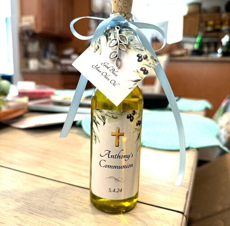 Puede incluir: Una peque&ntilde;a botella de vidrio llena de aceite de oliva, con tap&oacute;n de corcho y etiqueta decorativa. La etiqueta incluye una cruz dorada, el texto "Anthony's Communion" y la fecha 5.4.24. Una etiqueta con el texto "God Bless Your Olive Oil" est&aacute; atada con una cinta azul claro.