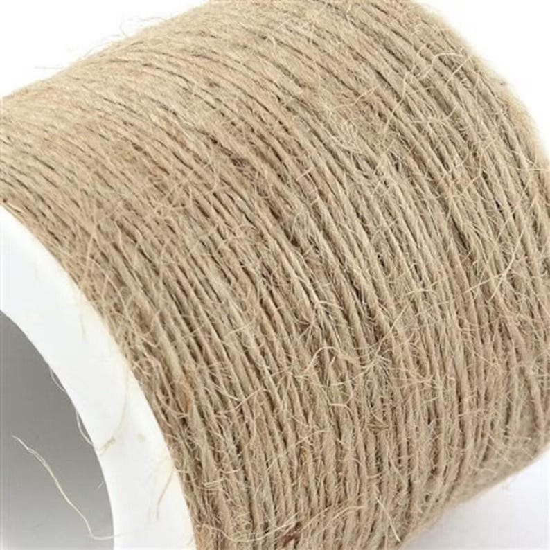 Puede incluir: Primer plano de una bobina de hilo de yute natural. El hilo es de color marr&oacute;n claro y est&aacute; enrollado firmemente alrededor de un n&uacute;cleo de cart&oacute;n blanco. La textura del hilo es visible, mostrando fibras individuales.