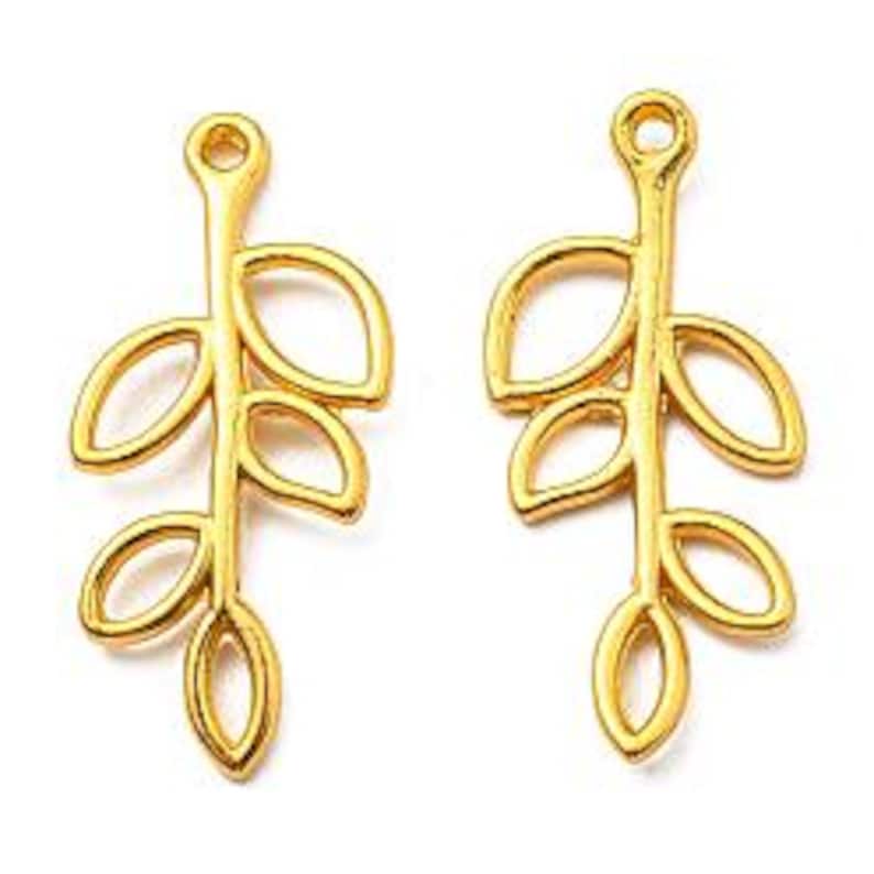 Puede incluir: Dos colgantes dorados con forma de hoja. Cada colgante presenta un tallo con tres hojas, cada una con forma ovalada. Los colgantes tienen un lazo en la parte superior para sujetarlos a la joyer&iacute;a. Los colgantes son de metal.